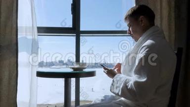 一个年轻有魅力的男人坐在<strong>靠窗</strong>的<strong>桌子</strong>旁喝茶，喝咖啡，在手机上写短信